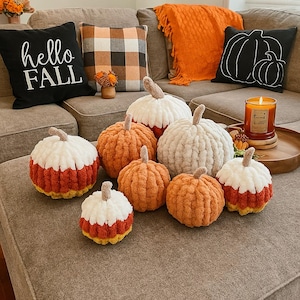May include: A cozy autumn scene featuring a collection of handmade, plush pumpkins in various sizes and colors. The pumpkins are orange, white, and candy corn-inspired, arranged on a neutral-toned sofa with decorative pillows and a candle.