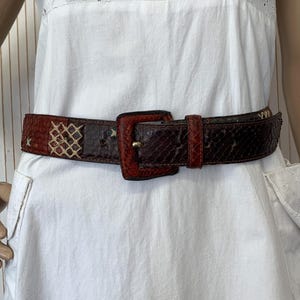 Peut inclure: Une ceinture en peau de serpent marron et rouge avec une boucle carrée. La ceinture présente un motif avec un mélange de couleurs, dont le rouge, le marron et le beige. La ceinture est portée sur une robe blanche.
