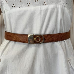 Peut inclure: Une ceinture en cuir marron avec une boucle argentée est portée sur une robe blanche. La ceinture présente un motif texturé. La robe a un corsage en œillet blanc et une jupe simple avec des poches.