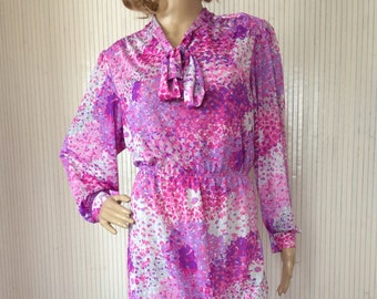 May include: A vintage midi dress in shades of pink, purple, and white, featuring a floral pattern. The dress has long sleeves, a tie-neck detail, and an elastic waist. The dress is displayed on a mannequin.