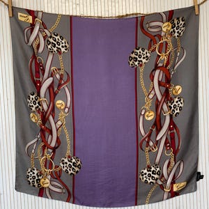 May include: A square silk scarf with a gray border and a central purple panel. The scarf features a pattern of red and white straps, gold chains, and leopard print accents. The brand name "Guess" is visible.