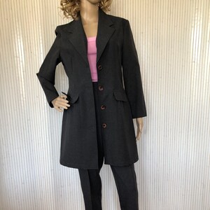 Könnte beinhalten: Ein dunkelgrauer Hosenanzug mit einem langen Blazer und einer geraden Hose. Der Blazer hat einen Reverskragen, einen Knopfschließe und Pattentaschen. Die Hose hat eine gerade Beinlinie und einen Knopfschließe.