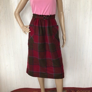 Peut inclure: Une jupe en tissu écossais rouge et marron avec une ceinture marron et deux poches. La jupe est mi-longue et a une taille froncée.