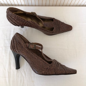 May include: A pair of brown leather heeled shoes with a woven pattern. The shoes have a t-strap design with a buckle closure.