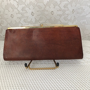 Puede incluir: Un bolso de mano rectangular marrón con un patrón texturizado similar al de un reptil. El bolso tiene un marco y un cierre de metal dorado. El bolso se exhibe sobre un soporte de metal dorado.