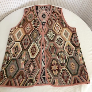 May include: A sleeveless vest with a geometric pattern in shades of pink, green, brown, and gold. The vest has a pink trim around the edges.