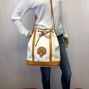 May include: A white canvas bucket bag with brown leather trim and a gold metal logo that reads "Trediadicous". The bag has a drawstring closure and a long brown leather strap.
