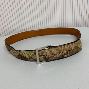 K&ouml;nnte beinhalten: Ein brauner und beigefarbener Schlangenleder-G&uuml;rtel mit einer silberfarbenen quadratischen Schnalle. Der G&uuml;rtel hat eine hellbraune Lederinnenseite und ein Patchwork-Design. Der G&uuml;rtel wird auf einer wei&szlig;en Oberfl&auml;che pr&auml;sentiert.