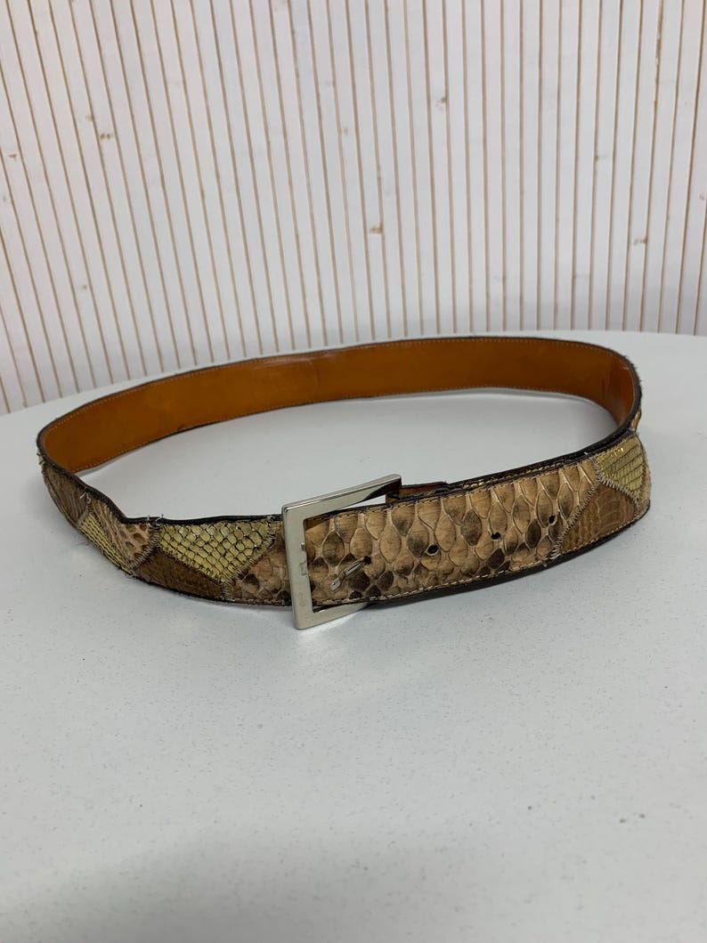 K&ouml;nnte beinhalten: Ein brauner und beigefarbener Schlangenleder-G&uuml;rtel mit einer silberfarbenen quadratischen Schnalle. Der G&uuml;rtel hat eine hellbraune Lederinnenseite und ein Patchwork-Design. Der G&uuml;rtel wird auf einer wei&szlig;en Oberfl&auml;che pr&auml;sentiert.