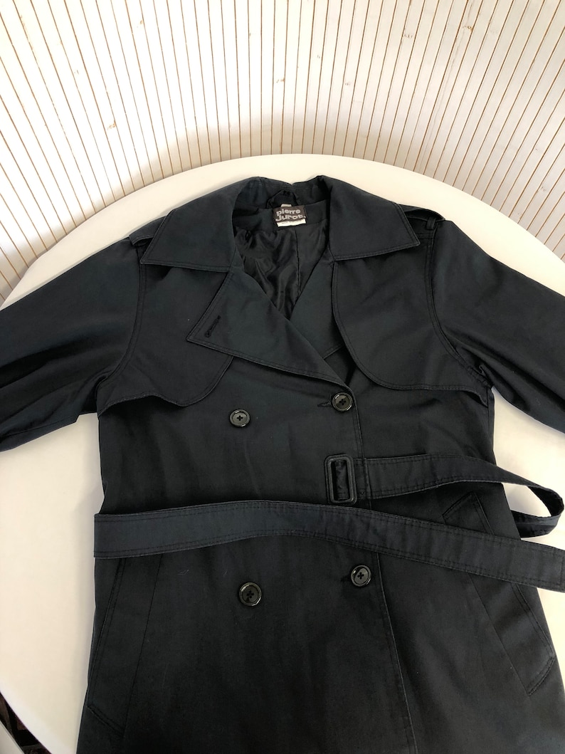 May include: A black double-breasted trench coat with a belt. The coat has a label that reads "Pierre Cardin" on the inside collar.