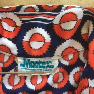 K&ouml;nnte beinhalten: Nahaufnahme eines Stoffes mit blauem, wei&szlig;em und orangefarbenem Muster und einem Etikett mit der Aufschrift "Mantex Creation Mantex Made in Belgium".