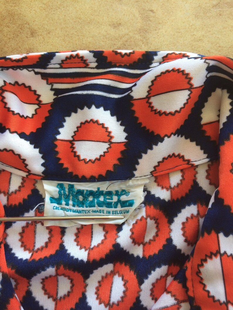 K&ouml;nnte beinhalten: Nahaufnahme eines Stoffes mit blauem, wei&szlig;em und orangefarbenem Muster und einem Etikett mit der Aufschrift "Mantex Creation Mantex Made in Belgium".