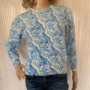 Puede incluir: Un suéter blanco con un patrón de paisley azul. El suéter tiene mangas largas y cuello redondo.