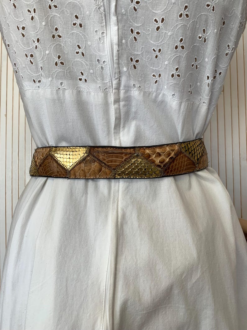 K&ouml;nnte beinhalten: Ein Vintage-G&uuml;rtel mit Schlangenmuster und goldenen Akzenten. Der G&uuml;rtel ist braun und goldfarben, mit einem geometrischen Design. Der G&uuml;rtel wird &uuml;ber einem wei&szlig;en Kleid mit Lochmuster getragen.