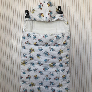 May include: White baby sleeping bag with a hood and a zip. The bag features a repeating pattern of blue, yellow, and red cartoon characters. The sleeping bag is suspended against a white wooden backdrop.