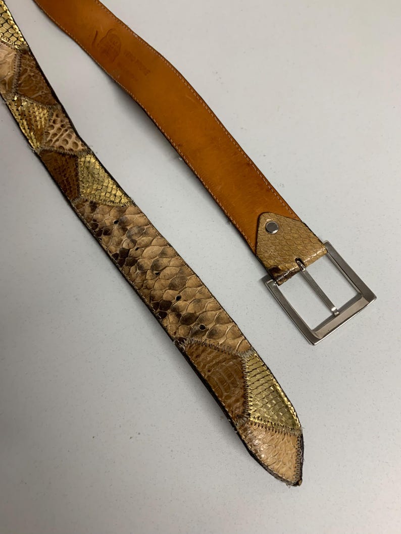 K&ouml;nnte beinhalten: Ein brauner Lederg&uuml;rtel mit Schlangenmuster. Der G&uuml;rtel hat eine silberne Schnalle und eine hellbraune Lederunterseite. Das Schlangenmuster weist Braun-, Gold- und Beiget&ouml;ne auf. Der G&uuml;rtel ist etwa 100-110 cm lang.