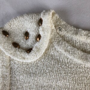 May include: A white sweater with a textured knit and a gold button trim around the neckline.