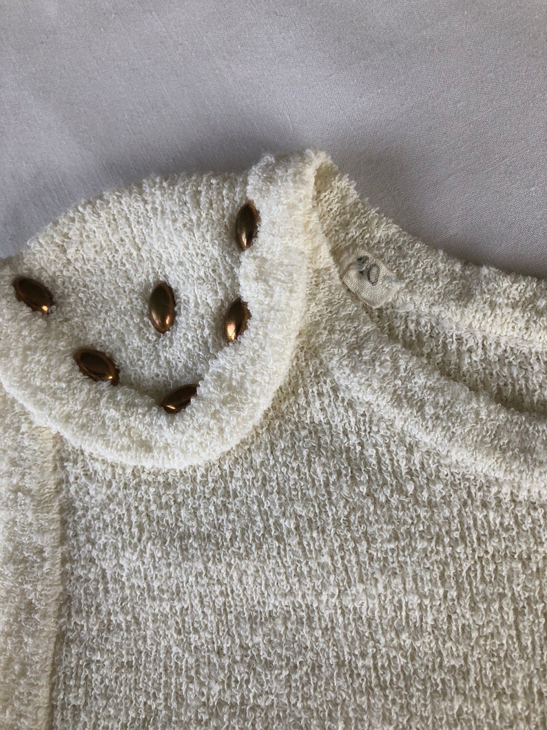 May include: A white sweater with a textured knit and a gold button trim around the neckline.