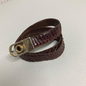 以下が含まれることがあります： ヘビ革模様の茶色のベルト。ベルトには金色のバックルが付いています。ベルトは白い表面に巻かれています。ベルトの幅は約2.5cmです。