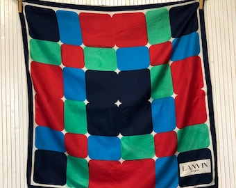 May include: A square silk scarf with a geometric pattern of red, blue, green, and navy blue squares. The scarf has a white border and the text "LANVIN PARIS" in the bottom right corner.