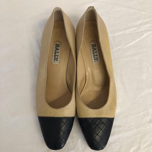 May include: A pair of beige leather pumps with black quilted toe caps. The shoes have a low heel and the brand name "BALLY" is visible inside each shoe. The shoes are displayed on a white surface.