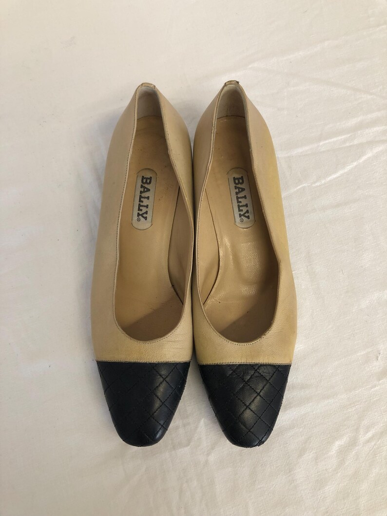 May include: A pair of beige leather pumps with black quilted toe caps. The shoes have a low heel and the brand name "BALLY" is visible inside each shoe. The shoes are displayed on a white surface.