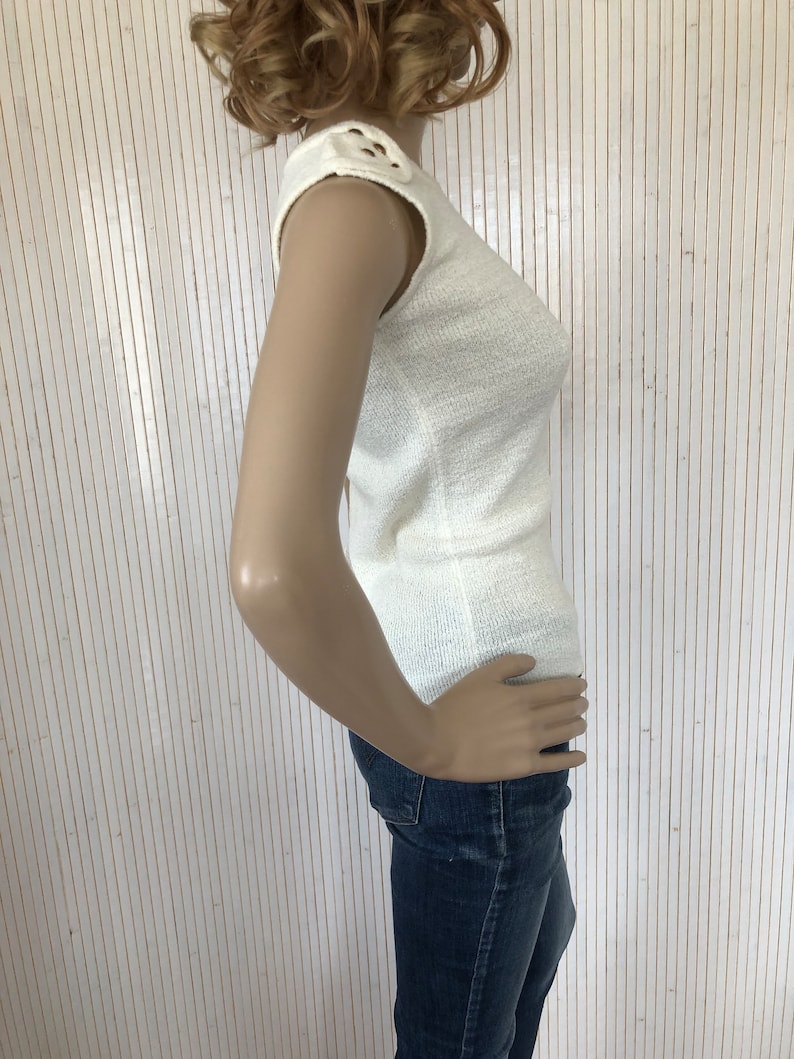 May include: A white sleeveless knit top with a decorative detail on the shoulder. The top is made of a soft, textured fabric.