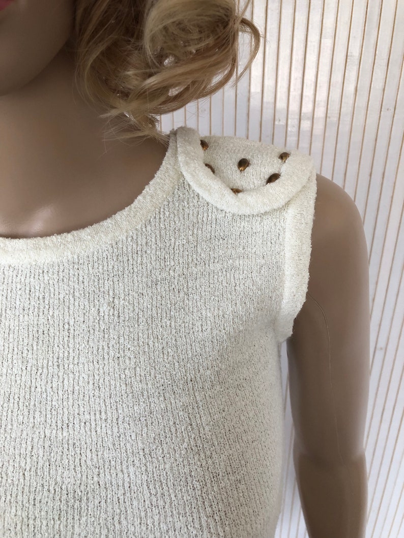 May include: White sleeveless top with a textured knit fabric and gold studs on one shoulder.