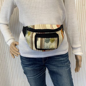 May include: A patterned fanny pack with a black zipper and a small front pocket. The bag features a cream-colored background with vertical stripes in shades of brown, red, and green. The fanny pack is worn around the waist.