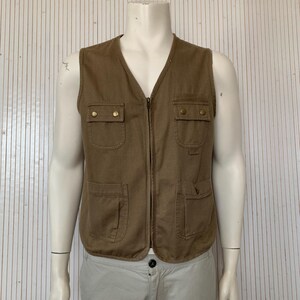 Könnte beinhalten: Eine braune Utility-Weste mit Reißverschluss und mehreren Taschen. Die Weste hat goldfarbene Knöpfe.