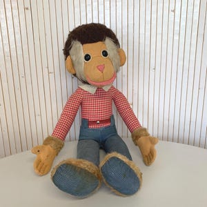 Op de afbeelding: Een vintage aapknuffel met een bruin hoofd, een beige gezicht en een roze mond. Het draagt een rood-wit geruit overhemd, een blauwe spijkerbroek en beige handschoenen en schoenen. De pop zit op een wit oppervlak.