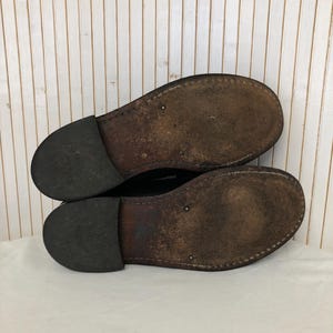 Pu&ograve; includere: Un paio di scarpe in pelle nera con suole in pelle marrone. Le scarpe sono usurate e hanno un aspetto vissuto.