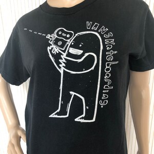 May include: Black t-shirt with a white graphic of a cartoon monster holding a video camera. The text "VANS Skateboarding." is printed on the shirt.