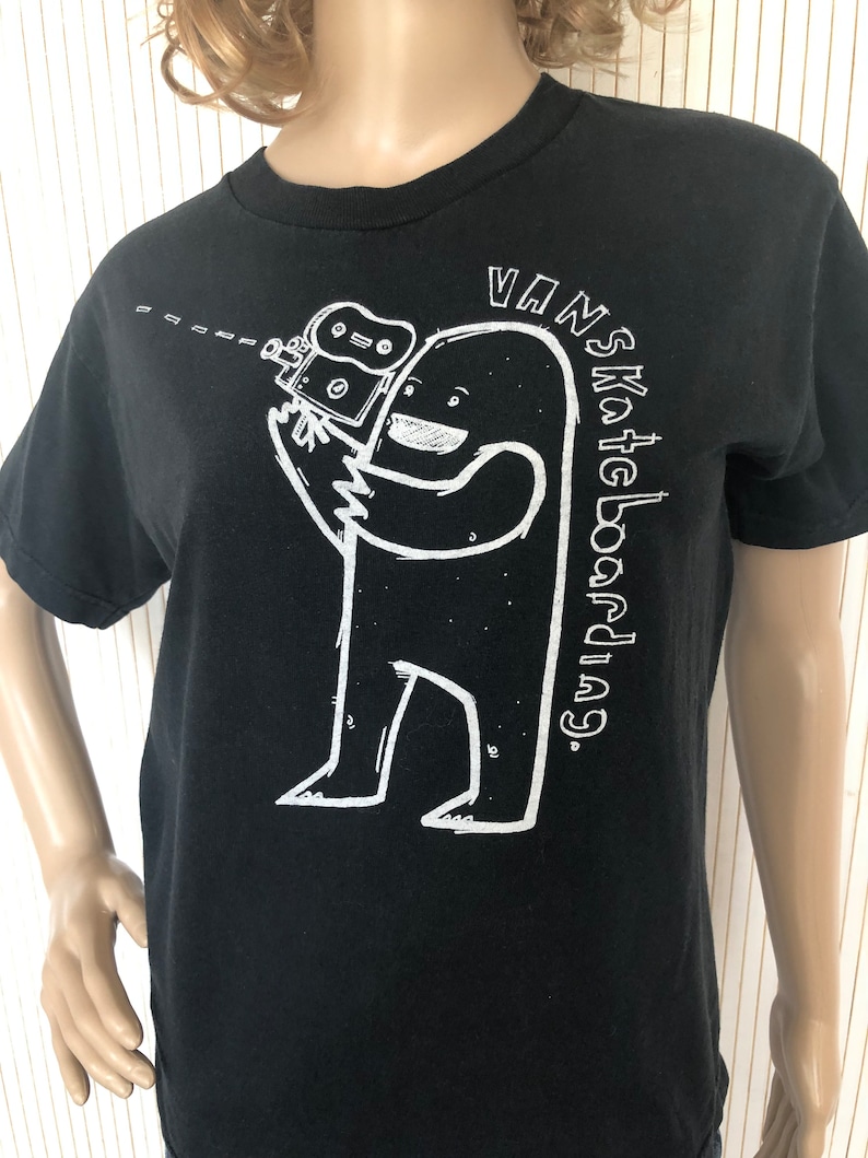 May include: Black t-shirt with a white graphic of a cartoon monster holding a video camera. The text "VANS Skateboarding." is printed on the shirt.