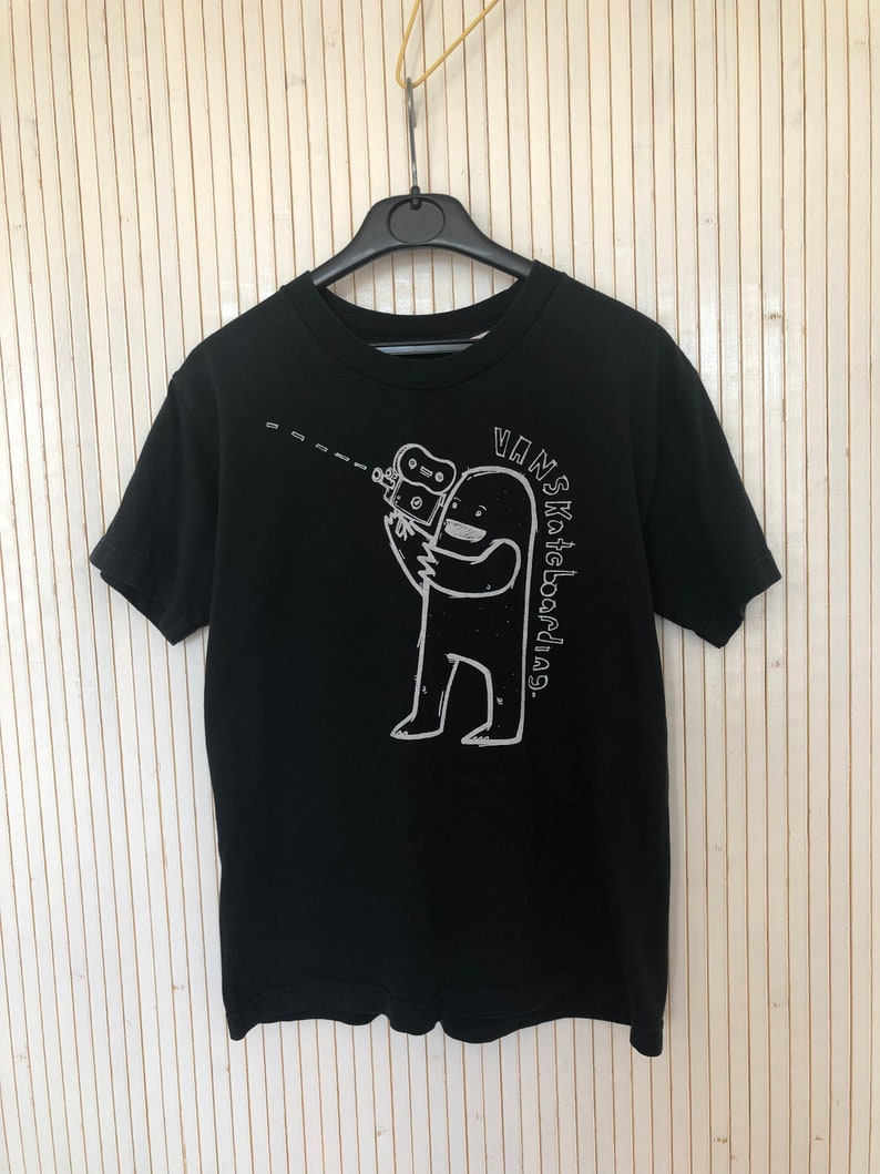 May include: Black t-shirt with a white graphic of a cartoon figure holding a camera and the text "Vans Skateboarding."