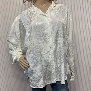May include: A cream-colored button-down shirt with a floral pattern. The long-sleeved shirt has a collar and cuffs, and is paired with blue jeans. The floral design is in a lighter shade of cream, creating a subtle contrast.