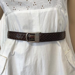 Op de afbeelding: Een bruine slangenleren riem met een zilverkleurige gesp wordt gedragen over een witte jurk. De riem is gemaakt van een textuurrijk materiaal en heeft een rechthoekige gesp. De jurk heeft een wit lijfje met broderie anglaise.