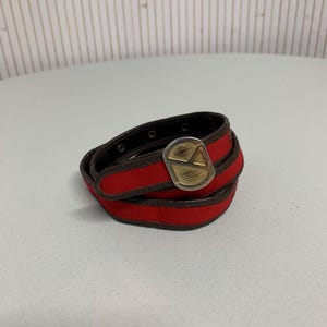 Könnte beinhalten: Ein aufgerollter roter Gürtel mit einer goldfarbenen Schnalle. Der Gürtel hat eine rote Stoffaußenseite und eine braune Lederinnenseite. Die Schnalle hat eine geometrische Form mit einem Dreieck-Design.