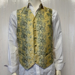 Può includere: Un gilet giallo e verde a fantasia floreale con chiusura a bottoni. Il gilet è indossato sopra una camicia bianca con colletto. Il gilet ha una lucentezza satinata e uno stile classico e formale.