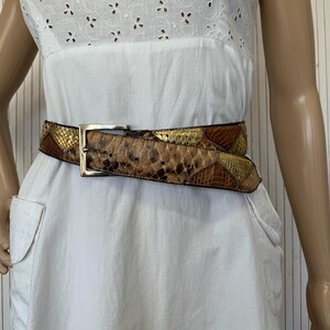 K&ouml;nnte beinhalten: Ein G&uuml;rtel mit Schlangenmuster und einer silberfarbenen Schnalle. Der G&uuml;rtel weist Farbt&ouml;ne von Braun, Gold und Beige auf. Der G&uuml;rtel wird &uuml;ber einem wei&szlig;en Kleid mit Lochmuster getragen.