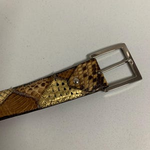 K&ouml;nnte beinhalten: Ein Schlangenleder-G&uuml;rtel mit silberner Schnalle. Der G&uuml;rtel hat ein Patchwork-Design in Braun-, Beige- und Goldt&ouml;nen. Die rechteckige Schnalle hat ein schlichtes, klassisches Design. Der G&uuml;rtel ist wahrscheinlich aus echtem Schlangenleder gefertigt.