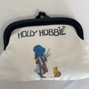 Op de afbeelding: Een vintage Holly Hobbie-portemonnee met een zwarte sluiting. De ecru stof heeft een print van het Holly Hobbie-personage, een meisje met een blauwe muts en jurk, met een kat. De tekst "HOLLY HOBBIE" staat boven de afbeelding.