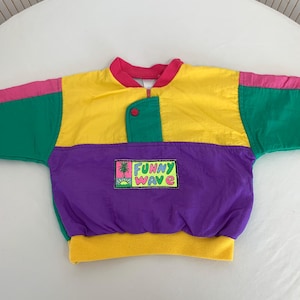 Peut inclure: Veste vintage colorée au design rétro. La partie supérieure est jaune, la partie inférieure violette, les manches vertes et les bordures roses. Le texte "FUNNY WAVE" et un motif tropical sont imprimés sur le devant.