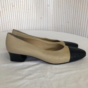 May include: A pair of beige leather flats with black quilted toe caps and black block heels. The shoes are displayed on a white surface against a backdrop of vertical wooden planks.