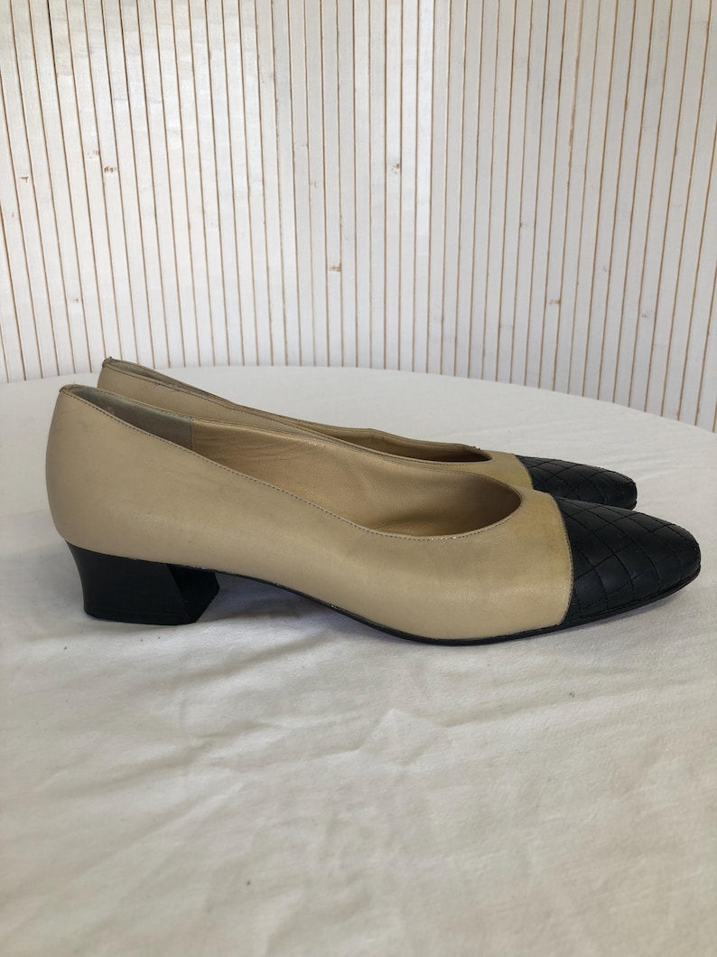 May include: A pair of beige leather flats with black quilted toe caps and black block heels. The shoes are displayed on a white surface against a backdrop of vertical wooden planks.