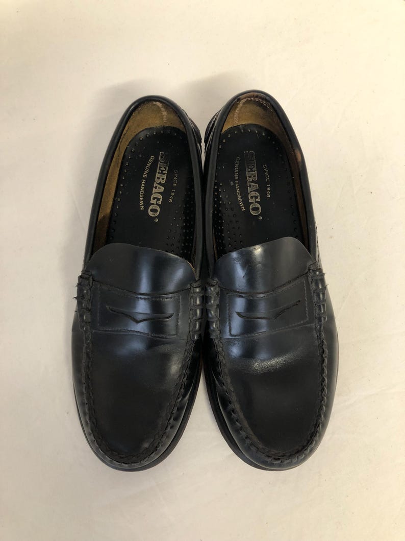 Pu&ograve; includere: Un paio di mocassini Sebago in pelle nera con un dettaglio di cinturino penny. I mocassini hanno un bordo cucito e una soletta interna in pelle marrone con il logo Sebago e "Since 1946" stampato su di essa.