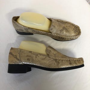 Op de afbeelding: Een paar beige slangenleren loafers met een lage hak. De loafers hebben een spitse neus en een slip-on ontwerp.