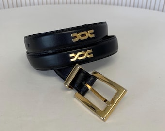 May include: Black leather belt with a gold-colored buckle and gold-colored decorative accents. The belt is rolled up on a white surface.