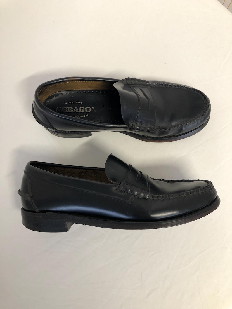Pu&ograve; includere: Un paio di mocassini in pelle nera con un dettaglio di cinturino penny. I mocassini hanno una soletta interna in pelle marrone con il testo "Sebago" e "Since 1946" stampato su di essa.