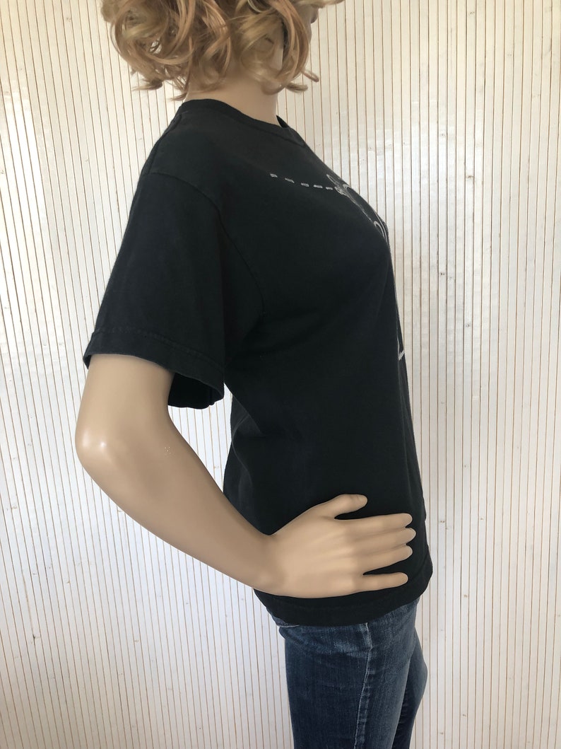 May include: A black short-sleeved t-shirt with a white graphic design. The design features a stylized image of a person with a microphone and a dashed line.